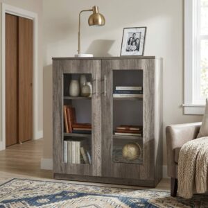 Mid-Height Wooden Credenza Bookcase With Textured Grey Wood-Grain Double Doors, Large Rectangular Glass Panels, Internal Storage Shelves, Long Silver Metal Handles, Flat Wooden Top, And Plinth Base.
